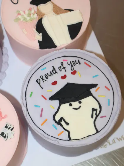 We love adding a touch of fun to our cakes! This "Proud of you" graduation bento cake features a cute, happy doodle character wearing a graduation cap, surrounded by colorful sprinkles.