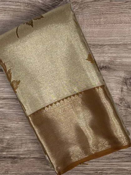 A shimmering gold tissue Kanchi silk saree with a brown border and delicate leaf motifs.