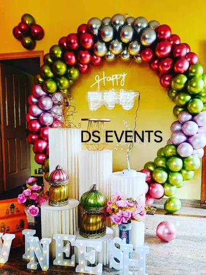 A vibrant and colorful setup using a metallic balloon arch in shades of pink, green, and silver. The decor is complemented by white plinths, marquee letters, and decorative lanterns for a festive look.