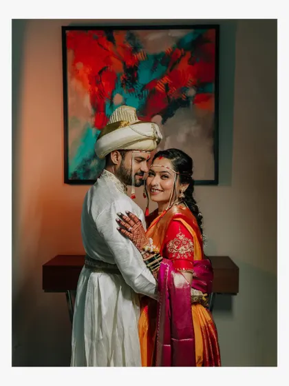 A romantic indoor portrait of a Maharashtrian couple. The groom in his white sherwani and the bride in her orange nauvari share an embrace in front of a modern art piece.