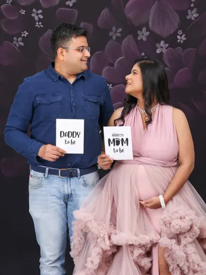 A classic studio announcement portrait. The couple sits together holding "Mom to be" and "Daddy to be" signs, looking at each other with love and excitement.