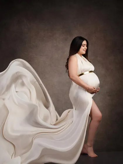 A dramatic fine art portrait featuring a white gown with a flowing train. The fabric is tossed to create a sense of movement against the dark, painterly backdrop.