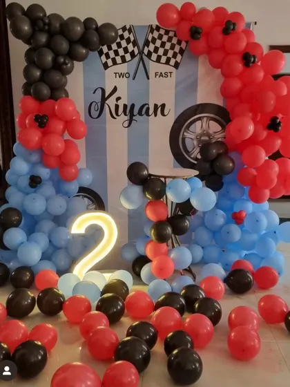 A "Two Fast" race car theme for a second birthday. The decoration includes a custom backdrop and balloon arches in red, blue, and black, with a light-up number '2'.