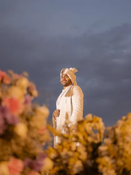 A stunning portrait of the groom against a dramatic twilight sky, framed by out-of-focus floral decor. This artistic shot highlights the groom's regal presence and the magical wedding atmosphere.