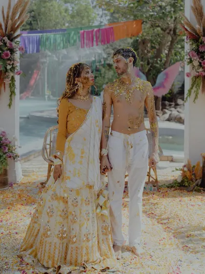 A post-Haldi portrait of the couple, holding hands amidst the scattered flower petals, their faces still showing the remnants of the joyful mess.
