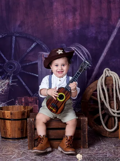 A happy little cowboy playing his guitar in a rustic barn setting. His big smile and the fun props create a playful and charming photo full of character.