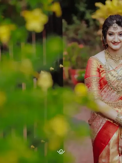 A portrait of a bride in a red and gold saree, framed by lush yellow flowers, creating a vibrant and natural setting.