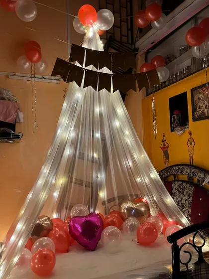 A cozy and intimate room decoration featuring a canopy of white net and fairy lights over the bed. The floor is covered with red, white, and pink balloons, perfect for a birthday or anniversary surprise.