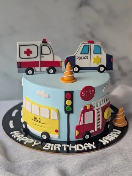 A cute transport-themed cake featuring a police car, ambulance, school bus, and fire truck. It's a colorful and fun design for a young vehicle enthusiast.