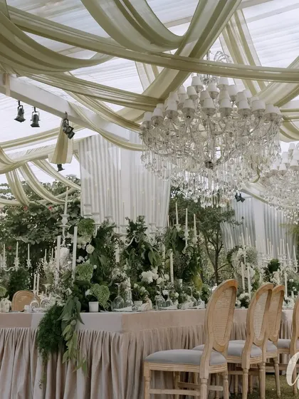 The main dining area under the tent, where flowing fabric and crystal chandeliers create a dreamy ceiling. The long tables are set with candelabras and lush greenery for a formal, elegant dinner.