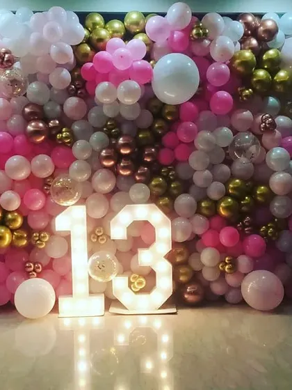 A fun and vibrant setup for a 13th birthday. A full wall of pink, white, and gold balloons creates a stunning backdrop for the light-up marquee '13', marking the official start of the teenage years.