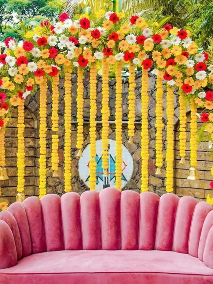 A vibrant and colorful setup for a baby shower. The decor features an arch of multi-colored gerberas and marigolds, with a modern pink sofa providing a comfortable and stylish seating area.