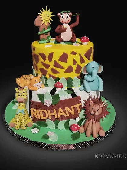 A two-tiered jungle animal cake for a first birthday. It features a giraffe-print top tier and is surrounded by adorable, hand-sculpted fondant animals like a lion, elephant, and tiger.