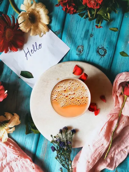 A vibrant flatlay with a cup of chai, colorful flowers, and a "Hello" note on a bright blue wooden background. This shot is cheerful, friendly, and full of personality.