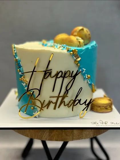 A stylish and modern birthday cake with a diagonal color block of white and teal buttercream. Gold sprinkles and a bold acrylic topper complete the look.