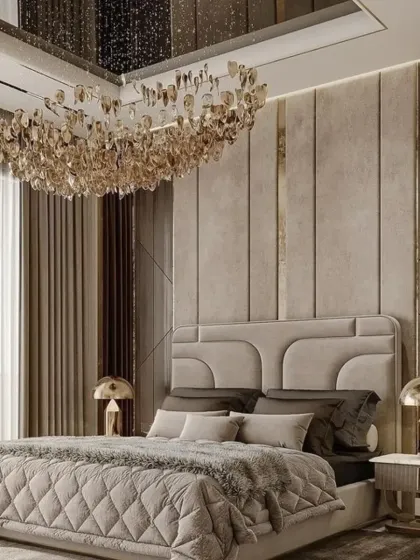 This bedroom achieves a soft, luxurious feel with a beige and gold color palette. The design includes a stunning crystal chandelier, a floor-to-ceiling padded headboard, and matching gold-accented table lamps.