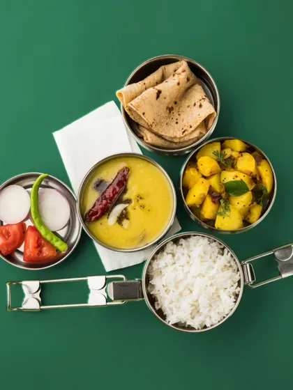 A classic steel tiffin, deconstructed to show the delicious contents: dal tadka, aloo sabzi, rice, and roti. This is the quintessential Indian lunch box, delivered.