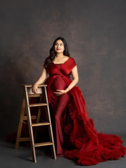 A classic studio portrait of an expecting mother in a rich red gown. She stands next to a wooden ladder prop, looking off to the side with a confident and happy expression.