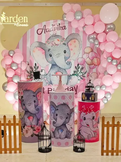 A sweet elephant-themed party in pink and grey. The setup includes a large circular backdrop, themed plinths, and a cute wooden fence.