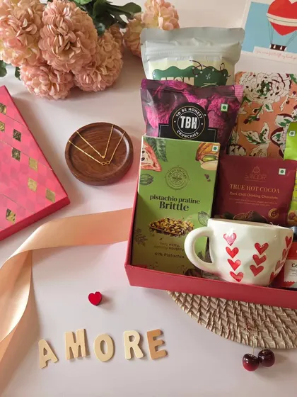 This "Amore" themed hamper is all about love. It features a heart-themed mug, pistachio praline brittle, and other gourmet treats, surrounded by flowers and a romantic ribbon.