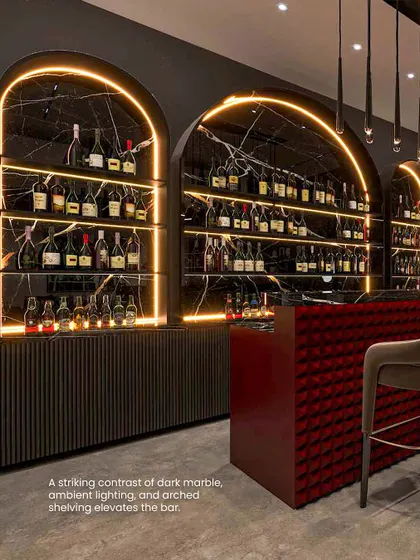 The bar is elevated by a striking contrast of dark marble, warm ambient lighting, and arched shelving. The red, tufted bar front adds a pop of color and texture.