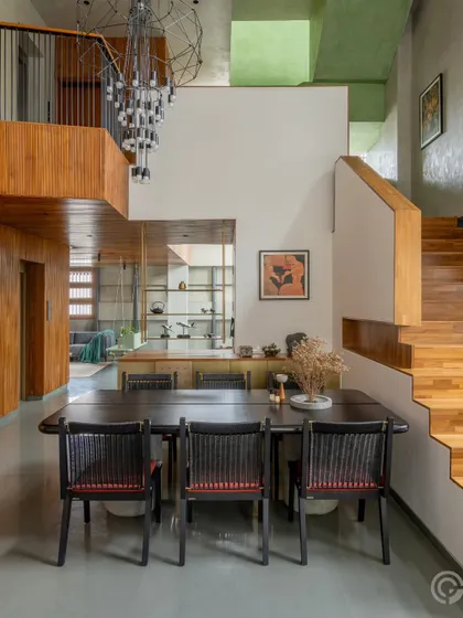 The dining area is situated within a double-height space, overlooked by a mezzanine level. The combination of warm wood paneling, a modern chandelier, and views into adjacent spaces creates a dramatic yet welcoming atmosphere for meals.