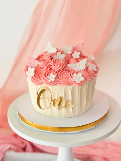 A giant cupcake cake for a first birthday. This design features pink rosette piping, a "One" topper, and delicate sugar flowers and butterflies.