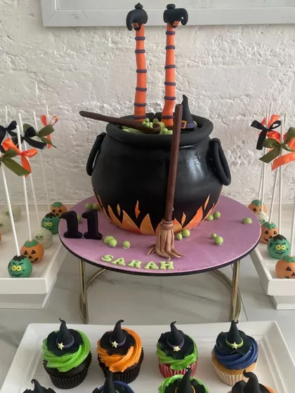 A spooky and fun Halloween dessert table. The centerpiece is a witch's cauldron cake, sculpted from cake and bubbling with green potion, with a witch's legs sticking out. The table also features witch hat cupcakes and pumpkin cakepops.
