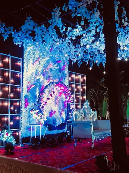 A dramatic stage design featuring a full wall of white flowers with pink accents, illuminated by cool blue lighting. The artificial tree with blue leaves adds a unique, artistic touch to the entire setup.