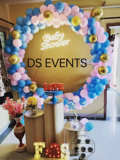 A simple yet beautiful baby shower setup perfect for home celebrations. The circular balloon arch in pink, blue, and gold frames the space, while the tiered stands are ready for the cake and gifts.