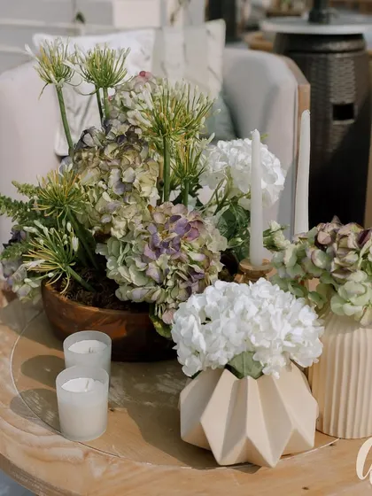 A close-up of the decor details, featuring muted green hydrangeas and white florals in modern ceramic vases. The natural wood table and simple candles complete the rustic yet elegant look.