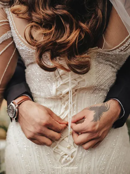 A close-up detail shot of the groom's hands lacing up the bride's dress. This intimate gesture is a beautiful symbol of their partnership and connection.