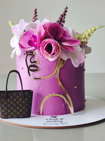 A fashion-forward birthday cake featuring a modern, abstract face outline in gold. The design is topped with a crown of fresh orchids and includes a miniature, edible designer handbag.