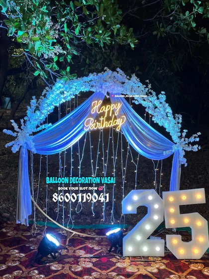 An outdoor birthday setup for a 25th celebration at night. The arch is decorated with white flowers and blue fabric, all illuminated with fairy lights and a "Happy Birthday" neon sign.