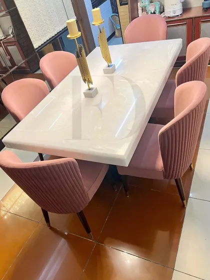 A six-seater dining set featuring a white onyx table and chairs upholstered in a soft pink velvet with vertical channel tufting. The tapered legs with gold caps add a final touch of elegance.