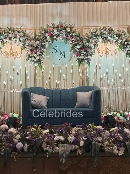 A beautiful sangeet stage setup with a deep blue sofa, candle-lit backdrop, and floral arrangements. The monogram adds a personal touch for the couple.