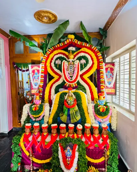 A goddess in a green saree inside a layered arch of red, yellow, white and green garlands, with a row of kalasha pots in front