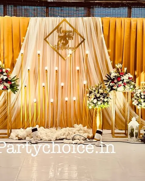 A hall stage with gold pleated drapes, a gold Happy 75th Birthday sign, tall gold candle stands and a lit 75 on the floor
