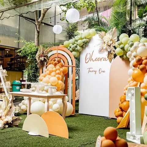 A first birthday set up in a garden cafe: a rainbow arch in peach and terracotta, a panel reading Turns One and a dessert table