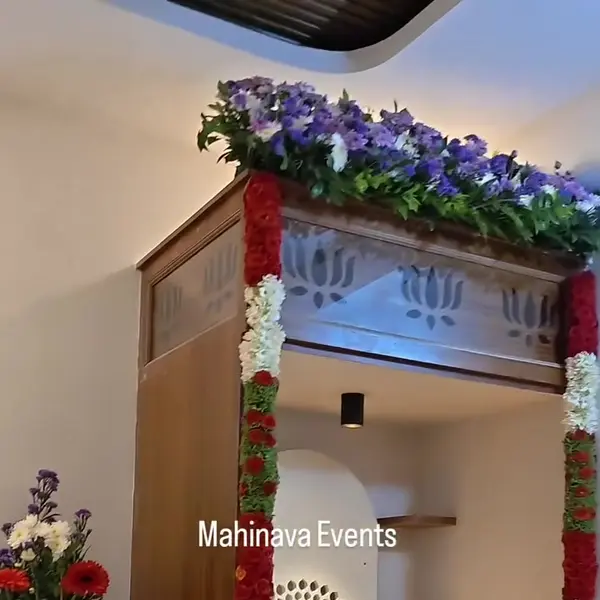 A wooden pooja niche in a new flat at a housewarming, framed in red and white garlands, with a crown of blue and white flowers on top and marigold petals on the shelves