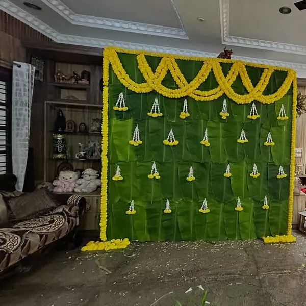 A wall of banana leaves in the hall of a house, with a scalloped swag of marigold across the top and small jasmine and marigold drops, beside a carved wooden door