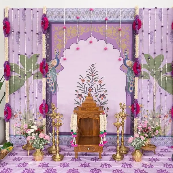 A lavender Pichwai panel with peacocks and a scalloped arch, flower strings with pink and purple flowers down both sides, a small wooden mandir in the middle, four tall brass lamps, pots of pink flowers and a lotus-print floor sheet