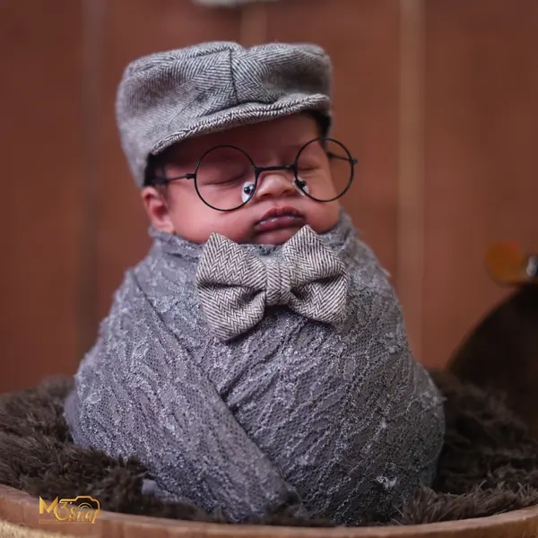 A newborn upright in a wooden bucket on brown fur, in a grey knit wrap, a flat cap, round glasses and a bow tie