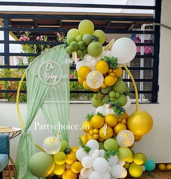 On a balcony: a gold ring with a sage drape and a white Oh Baby neon sign, a garland of sage, mustard, white and clear confetti balloons with leaves, and a small gold table