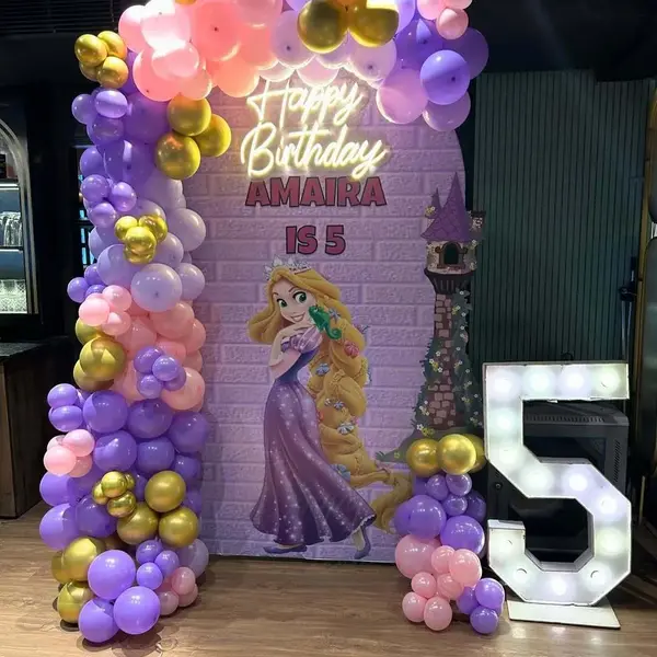 A fifth birthday: an arched purple brick-print backdrop with Rapunzel and her tower, reading Happy Birthday Amaira Is 5 under a warm Happy Birthday neon sign, a purple, pink and gold balloon garland, and a lit 5 beside it