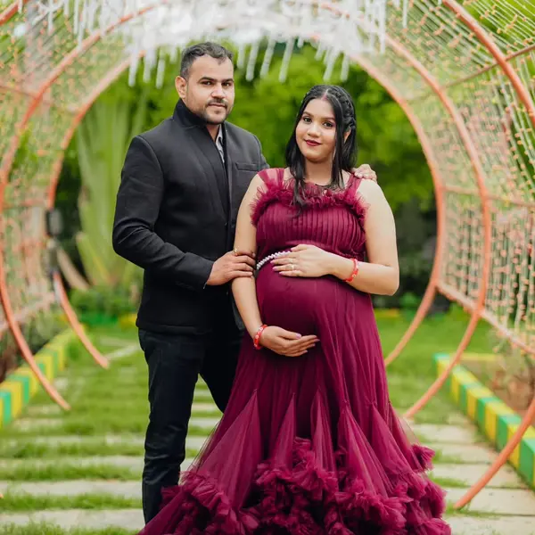 A pregnant woman in a wine tulle gown stands with her husband in a dark suit under a lit arch on an outdoor set