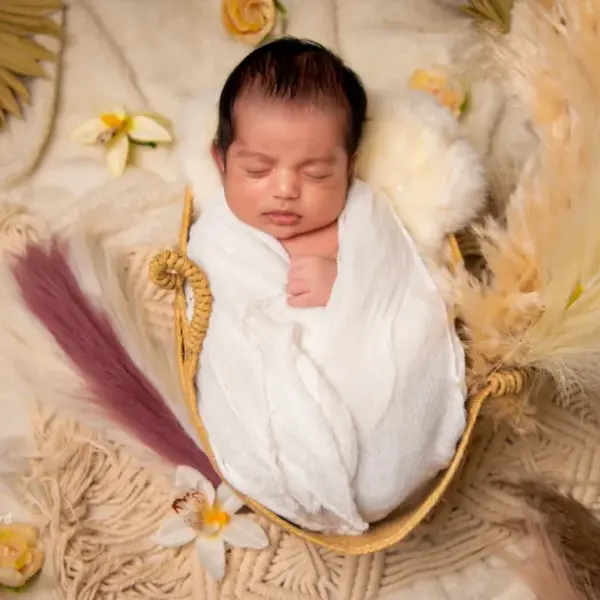 A newborn asleep in a white wrap in a small woven basket on a cream throw with pampas grass, a palm leaf and frangipani flowers