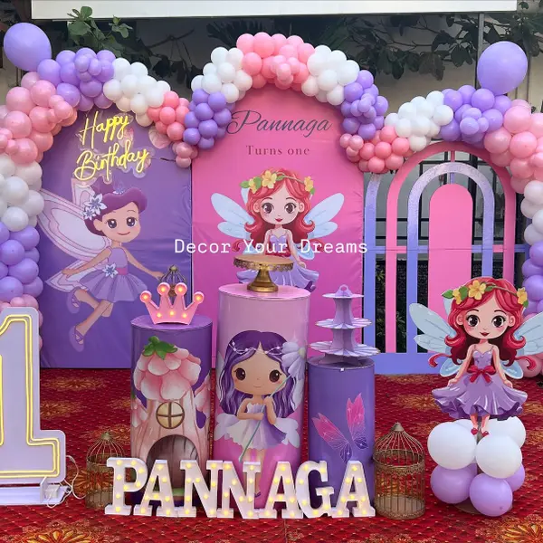 A fairy first birthday outdoors: three printed arched panels with fairy art, the name and Turns one, a pink, lilac and white balloon garland over them, themed plinths, a standing fairy cutout, a lit 1 and the name in lit letters on a red carpet