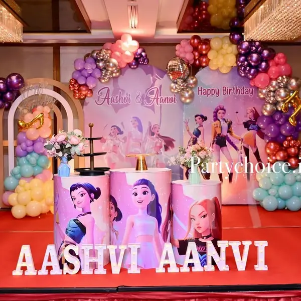 A K-pop theme stage on a red carpet: two printed panels and three plinths with an illustrated girl group, a purple, pink and gold balloon arch, disco balls and two names in white letters