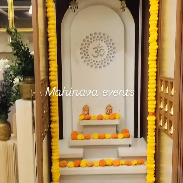 A white pooja unit with an Om panel in a flat, framed in long strings of yellow marigold, with a thick marigold header, the edge of a red rose top above it, small bells, and marigold heads on the shelves